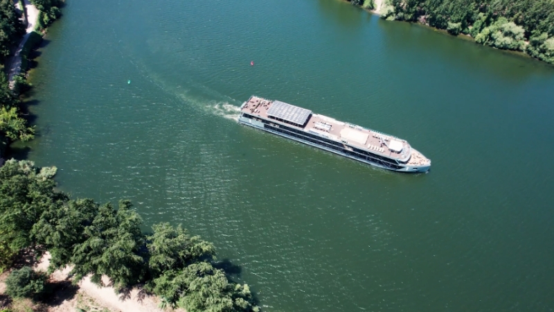 A river cruise ship moves through calm green water near a forested shoreline